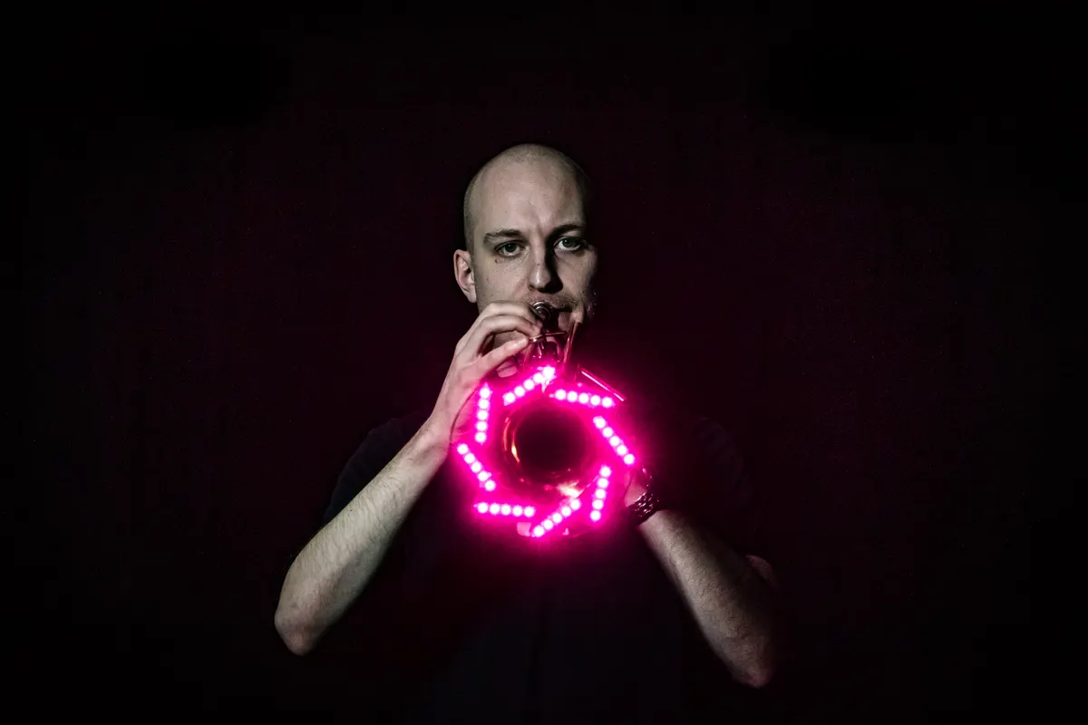 Brass player holds a pink LED-lit instrument on a dark stage.