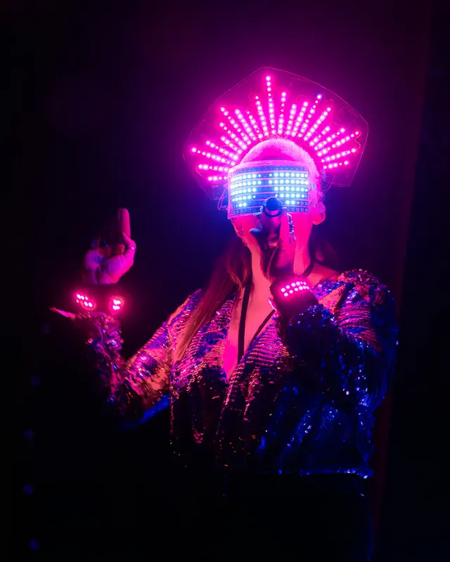 Vocalist in a glowing LED headdress and visor performs on a dark stage.