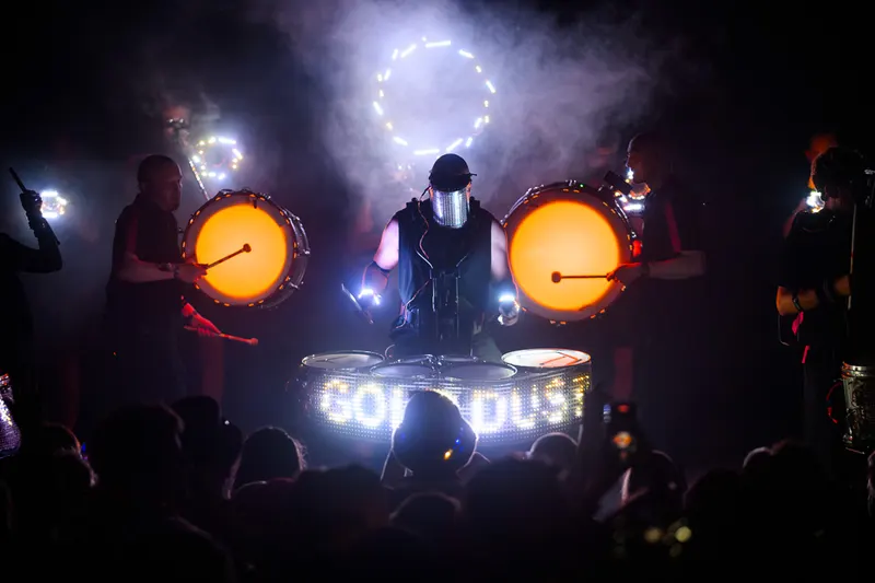 Drummers play glowing orange drums through stage smoke.