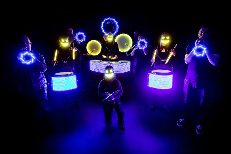 Band members pose with bright LED-lit brass instruments and drums.