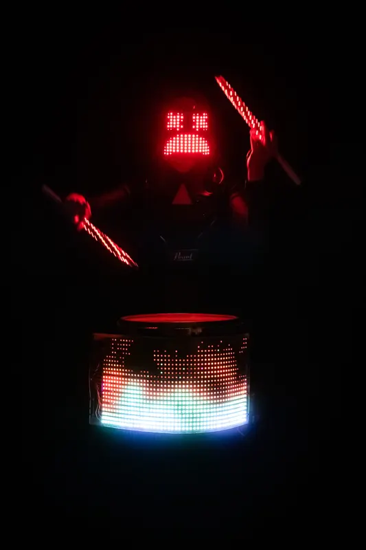 Drummer in a red LED mask raises glowing sticks above an illuminated drum.